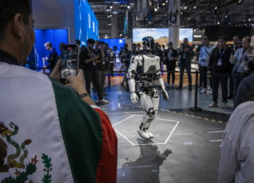 Visitante graba a un robot humanoide en una feria tecnológica, con asistentes alrededor y una persona con referencia mexicana en su ropa.